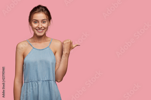 Studio shot of pretty Caucasian woman with glad facial expression, points wit...
