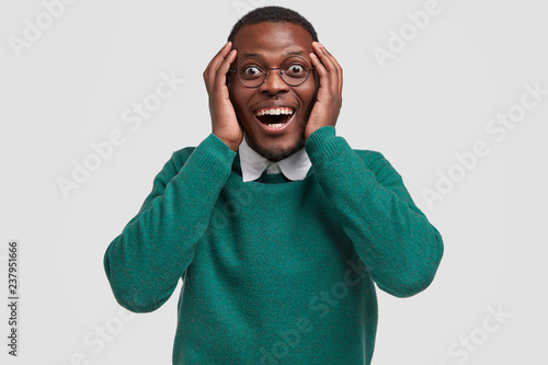 Positive young African American guy feels overjoyed as recieves money reward for good diligent job, keeps hands on head, smiles broadly, wears casual green jumper, isolated over white background
