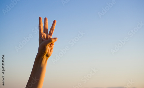 Hand doing / showing number three gesture symbol on blue summer sky nature background. Gesturing number 3. Number three in sign language. Third and counting down three concept. Three fingers up.