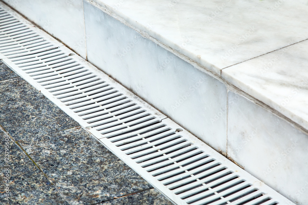 Obraz premium The gutter of the drainage system on the sidewalk, close-up metal grille light color on the background of marble tiles.