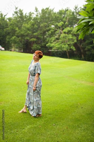 portrait of carefree adult caucasian redhead woman in dress posing in green summer park.