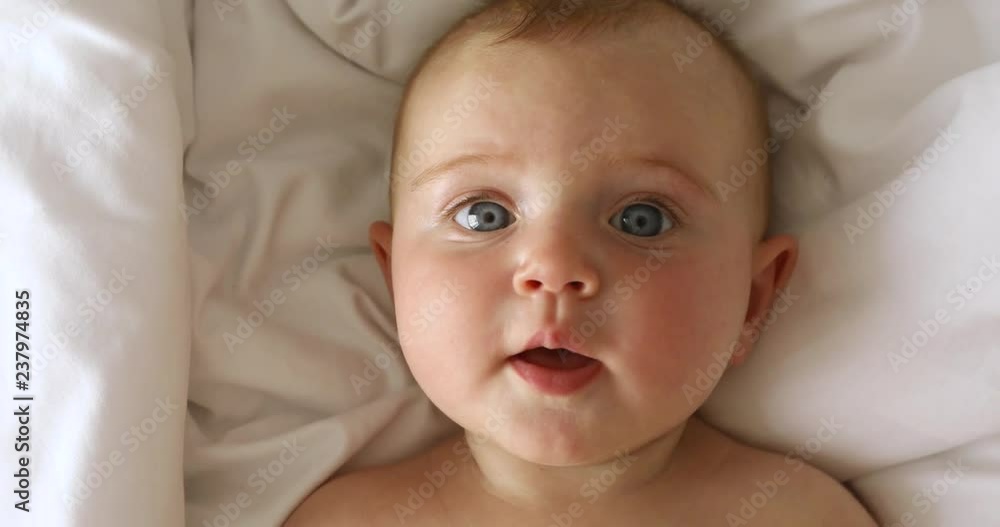 Cute little baby is looking into the camera and is happy on a white bed sheet Cute little baby is looking into the camera and is happy on a white bed sheet