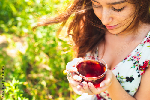 Young beautiful girl with long eye lashes with glass cup of black tea, happy teen girl smiling in floral summer dress drinking healthy herbal hot drink in garden, green nature, copy space