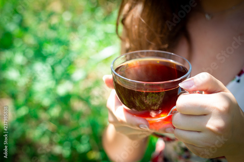 Young beautiful girl with long eye lashes with glass cup of black tea, happy teen girl smiling in floral summer dress drinking healthy herbal hot drink in garden, green nature, copy space