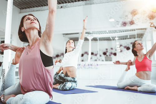 Close-up view on strong healthy woman practicing yoga exerciese on fitness mat in white sport class. Adult fit female doing stretching early morning indoor on background people