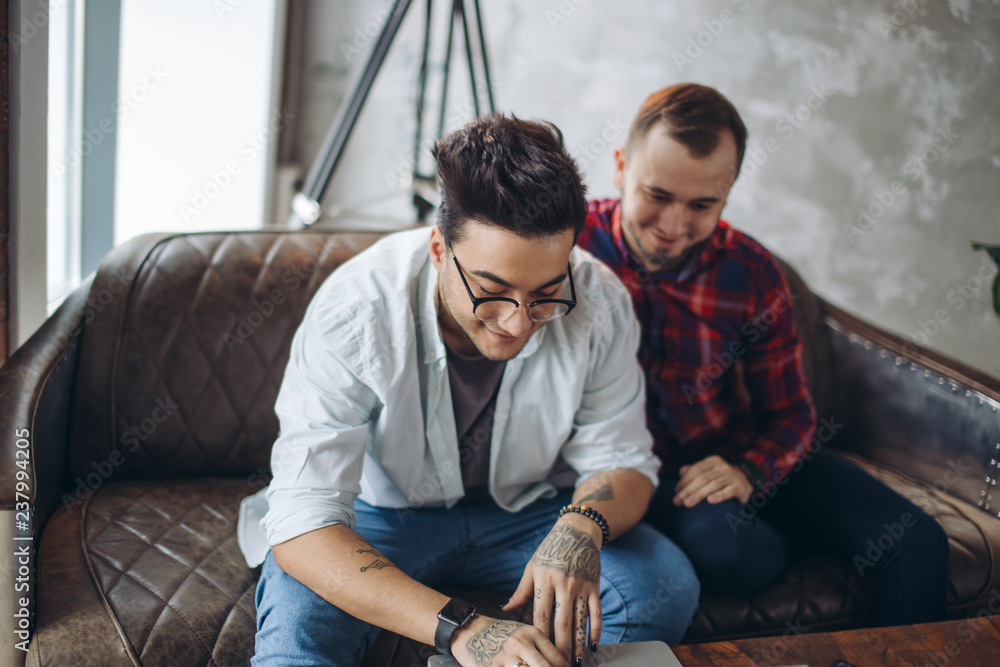 Romantic partners in love and business. Caucasian amused gay couple looking at laptop screen while resting in living room with loft interior. Dominant man wearing spectacles and and using laptop