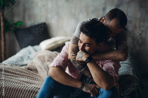 Happy gay couple embraced, joking and having fun in an intimate hug. Positive blue man embraces his friend, happy to be close to lover, sitting on bed at home, close up. Same-sex love relationship. photo