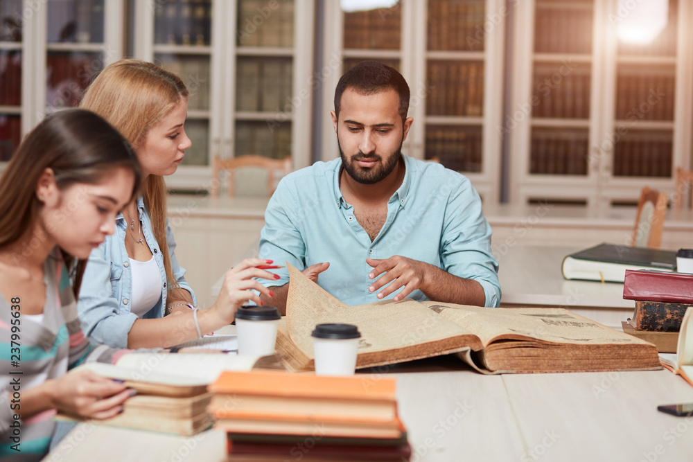 Multiracial students learning in college library, sitting at the table with big old reference textbook, writing down and discussing notes. Multiracial students learning in college library, sitting at the table with big old reference textbook, writing down and discussing notes.
