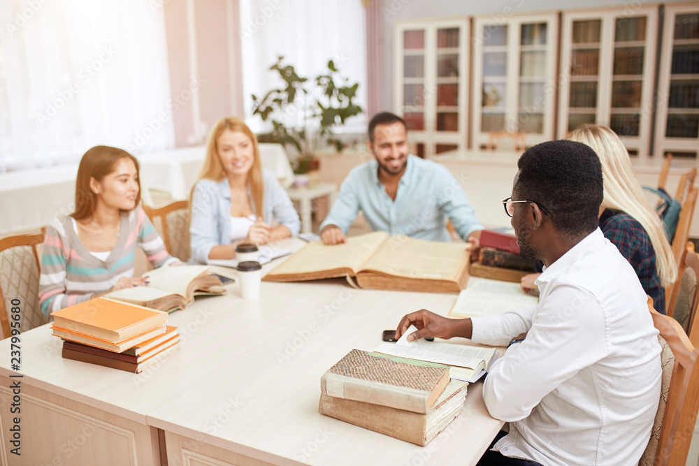 Multiracial students learning in college library, sitting at the table with big old reference textbook, writing down and discussing notes. Multiracial students learning in college library, sitting at the table with big old reference textbook, writing down and discussing notes.