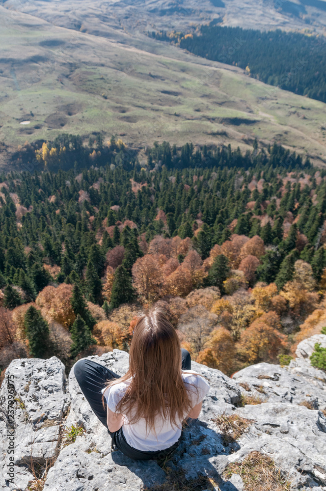 a young active girl sits on the edge of a rock, look at the Caucasus mountains.