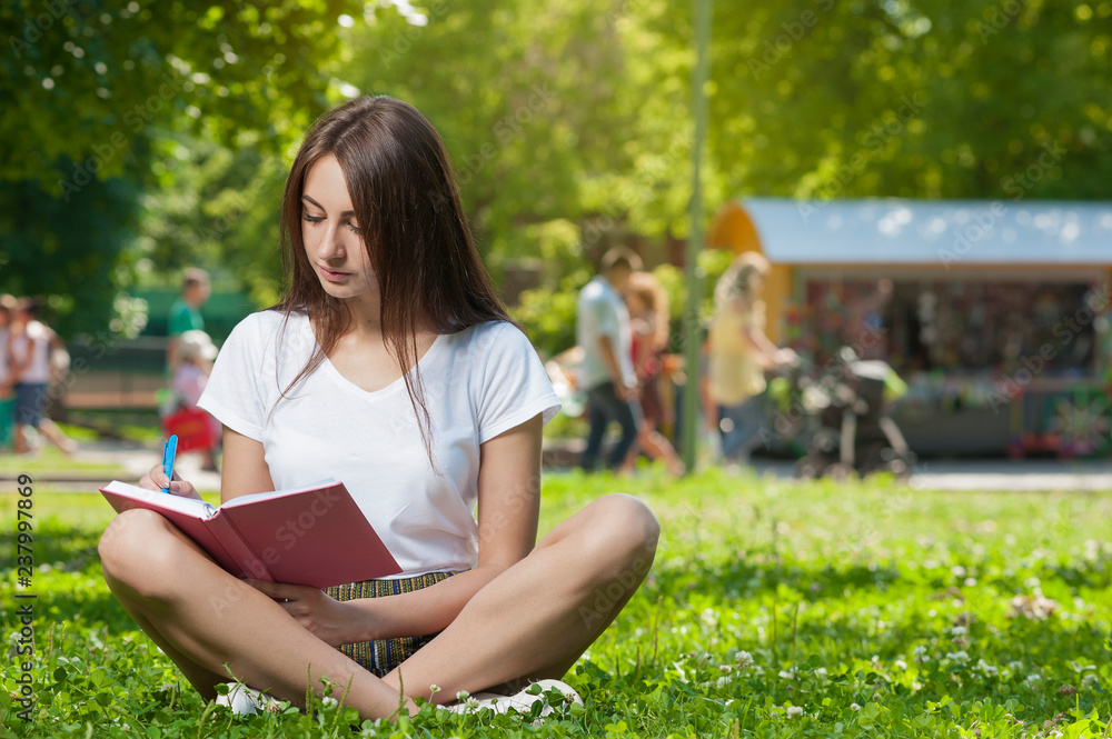 Obraz premium Brunette Student Girl Sitting on Green Grass in Park