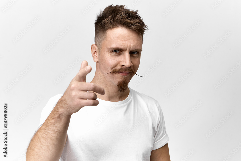 Signs, symbols, gestures and body language concept. Horizontal view of confident joyful young handsome guy with trendy hairstyle, mustache and beard smiling and pointing fore finger at camera Signs, symbols, gestures and body language concept. Horizontal view of confident joyful young handsome guy with trendy hairstyle, mustache and beard smiling and pointing fore finger at camera