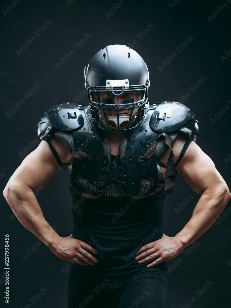 Portrait of sexy american football player wearing protective armour on shirtless torso, looking confidently, ready to tear apart everybody who bothers to win, isolated over dark studio background.