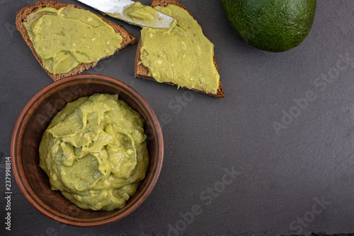 Fresh avocado cream in brown bowl with bread slices on black background. Top view. With copy space photo