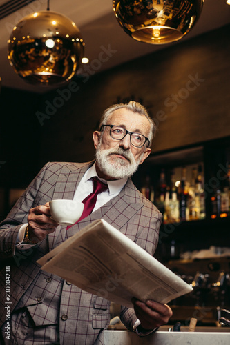 Grey haired old wealthy man, the famous publisher dressed in formal three - pieces suit reading newspaper while having morning cup of coffee at his favourite coffee shop near his office