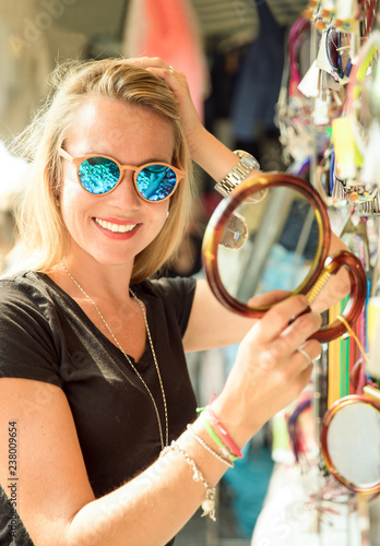 junge hübsche blonde frau auf dem markt in der sommer sonne in italien bei ihrer auslands reise. sie schaut sich sonnenbrillen, t-shirts, souveniers und taschen am markstand an und  photo