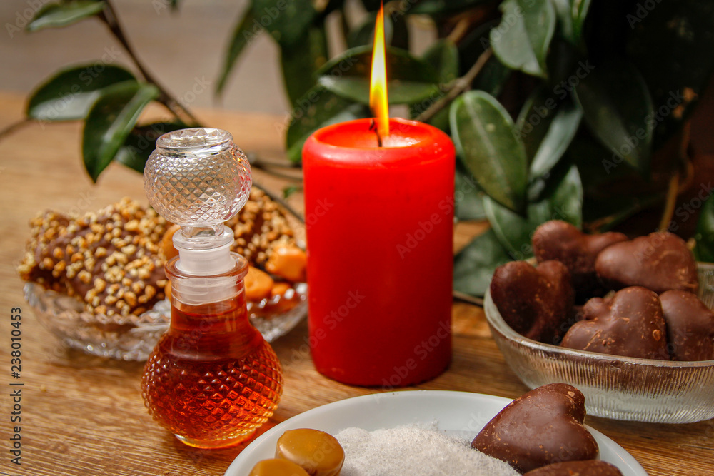Bottle of perfume and various of cookies on wooden table with red candle. Home liana in the background. Close up. Bottle of perfume and various of cookies on wooden table with red candle. Home liana in the background. Close up.