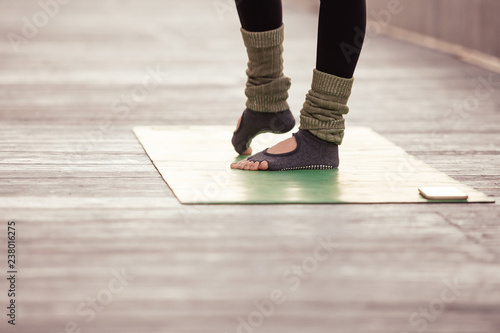 Young yogi instructor doing a leg workout before yoga class. Woman wearing sportswear green pants, standing on mat outdoor at yoga wooden terrace in the morning. Healthy hobby well being concept