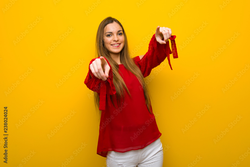 Young girl with red dress over yellow wall points finger at you while smiling Young girl with red dress over yellow wall points finger at you while smiling