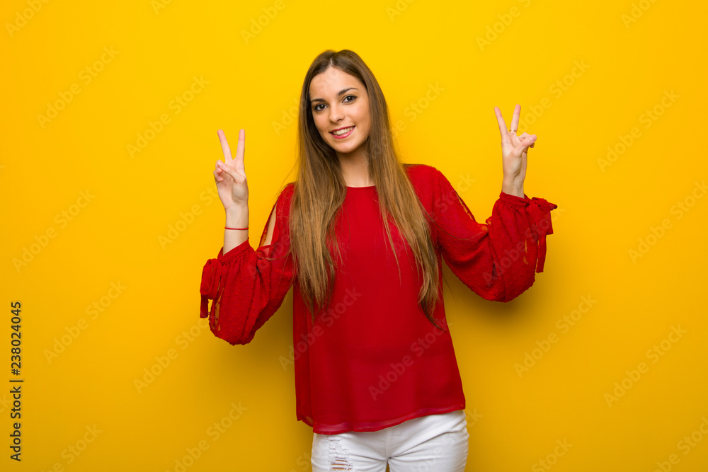 Young girl with red dress over yellow wall smiling and showing victory sign with both hands Young girl with red dress over yellow wall smiling and showing victory sign with both hands