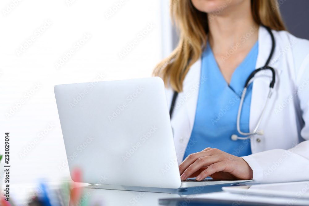 Doctor typing on laptop computer while sitting at the glass desk in hospital office. Physician at work. Medicine and healthcare concept Doctor typing on laptop computer while sitting at the glass desk in hospital office. Physician at work. Medicine and healthcare concept