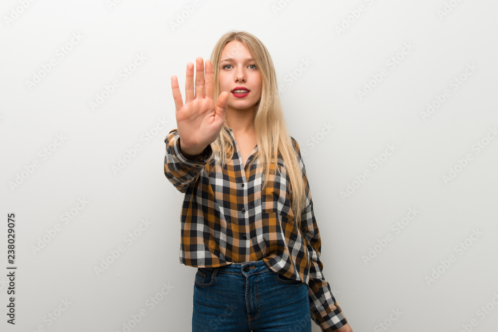 Blonde young girl over white wall making stop gesture denying a situation that thinks wrong Blonde young girl over white wall making stop gesture denying a situation that thinks wrong