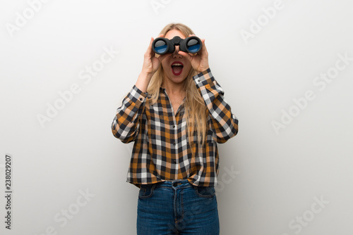 Blonde young girl over white wall and looking in the distance with binoculars