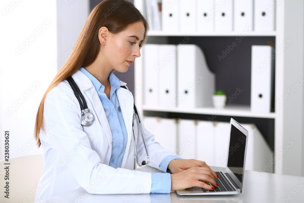 Doctor woman typing on laptop computer while sitting at the desk in hospital office. Physician at work. Medicine and health care concept Doctor woman typing on laptop computer while sitting at the desk in hospital office. Physician at work. Medicine and health care concept