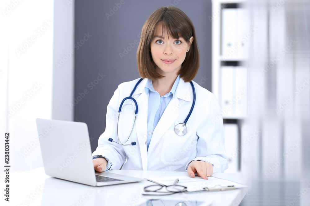 Doctor typing on laptop computer while sitting at the white table in hospital office. Physician at work. Medicine and healthcare concept Doctor typing on laptop computer while sitting at the white table in hospital office. Physician at work. Medicine and healthcare concept