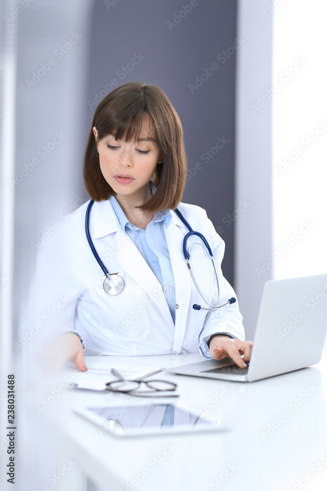 Doctor typing on laptop computer while sitting at the white table in hospital office. Physician at work. Medicine and healthcare concept Doctor typing on laptop computer while sitting at the white table in hospital office. Physician at work. Medicine and healthcare concept