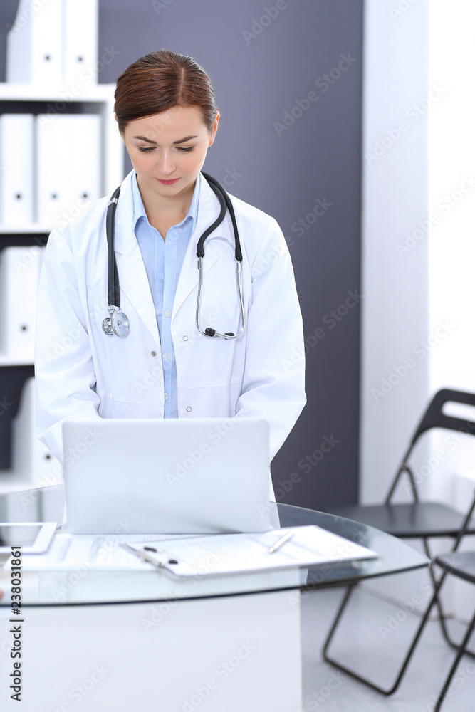 Happy doctor woman at work. Portrait of female physician using laptop computer while standing near reception desk at clinic or emergency hospital. Medicine concept Happy doctor woman at work. Portrait of female physician using laptop computer while standing near reception desk at clinic or emergency hospital. Medicine concept