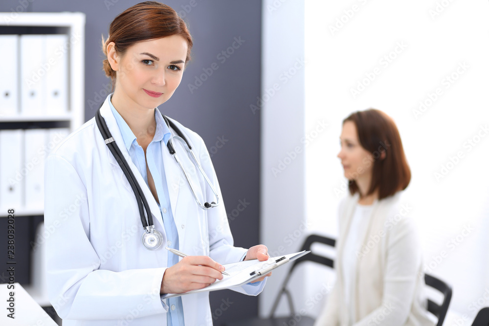 Doctor woman at work. Portrait of female physician filling up medical form while standing near reception desk at clinic or emergency hospital. Patient woman sitting at the background. Medicine concept Doctor woman at work. Portrait of female physician filling up medical form while standing near reception desk at clinic or emergency hospital. Patient woman sitting at the background. Medicine concept