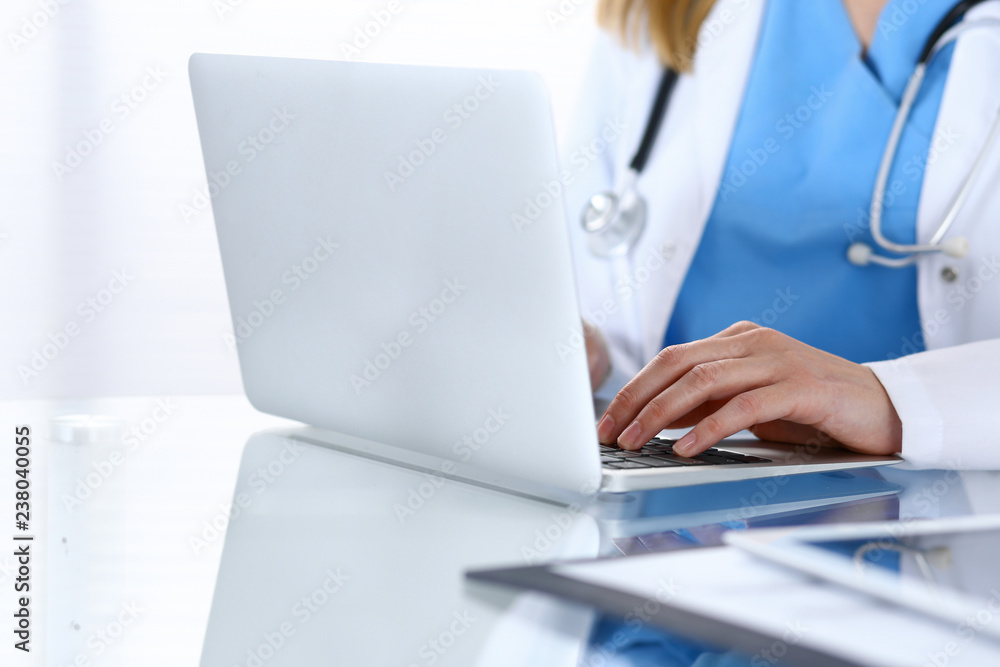 Doctor typing on laptop computer while sitting at the glass desk in hospital office. Physician at work. Medicine and healthcare concept Doctor typing on laptop computer while sitting at the glass desk in hospital office. Physician at work. Medicine and healthcare concept