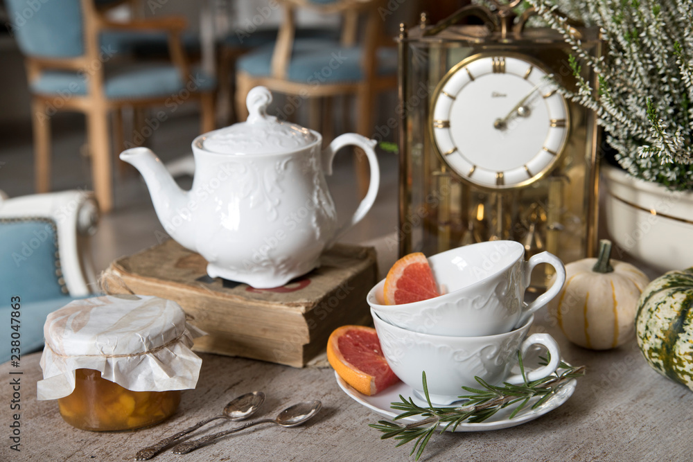 Fototapeta premium Still life. White beautiful teapot, cups and saucer, apricot jam, grapefruit and rosemary. Against the backdrop of vintage watches and heather.