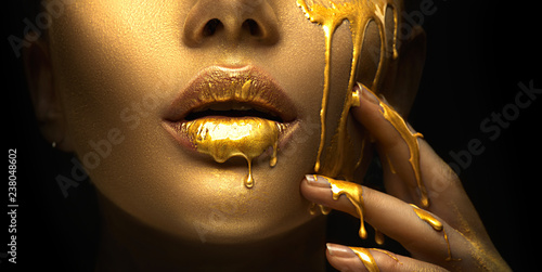 Golden paint smudges drips from the face lips and hand, golden liquid drops on beautiful model girl's mouth, creative abstract makeup. Beauty woman face
