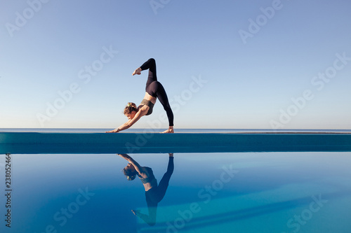 Young attractive woman practicing yoga stand doing Downward facing dog exercise, adho mukha svanasana pose, working out by the pool, above beach, relaxing against blue sky. Health and beauty concept © Rithor