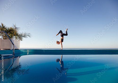 Young attractive woman practicing yoga stand in Adho Mukha Vrksasana exercise, Downward facing Tree pose, working out by the pool, above the beach, relaxing against blue sky. Health and beauty concept © Rithor