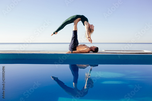 Acro yoga concept. Pair yoga. Couple of young sporty people practicing yoga lesson with partner, man and woman in yogi exercise, arm balance pose, working out by pool, above beach, against blue sky photo