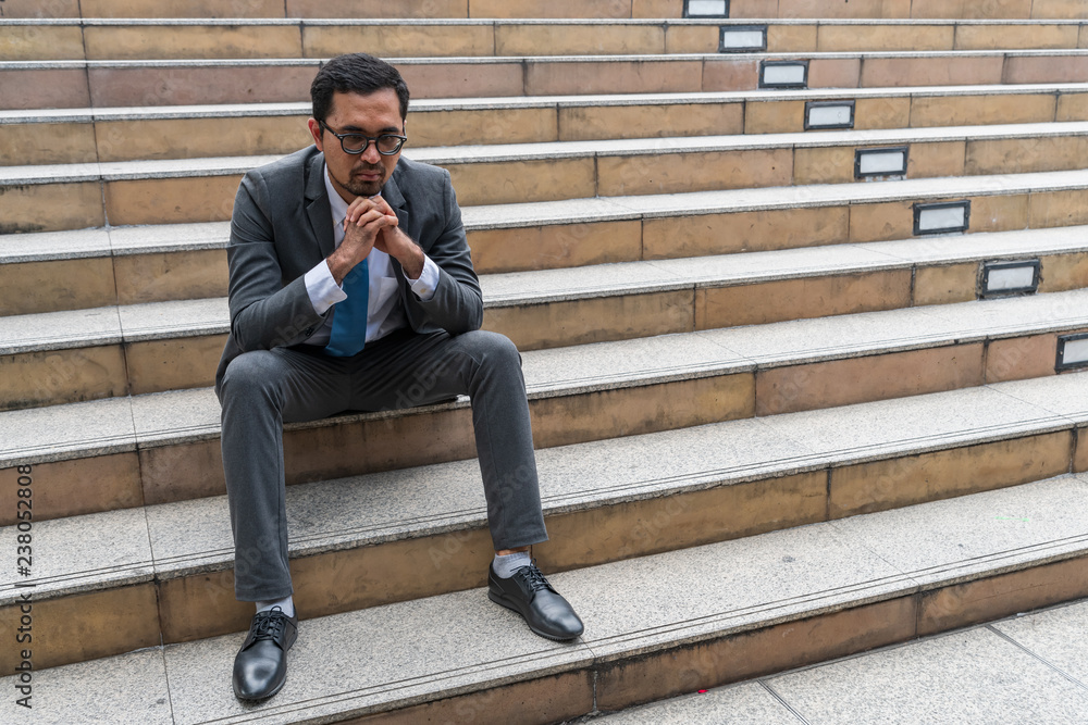 A sad looking young businessman sitting at the staircase looking down with stressed and depressed look gesture A sad looking young businessman sitting at the staircase looking down with stressed and depressed look gesture