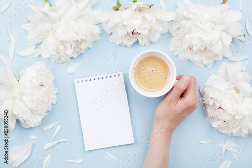 Woman hand with cup of coffee, open notebook and beautiful white peony flowers on pastel table top view. Cozy breakfast in flat lay style.