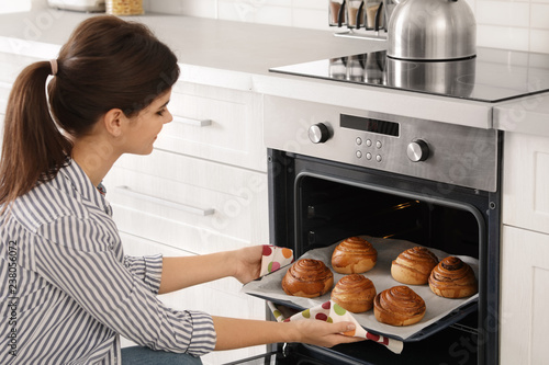Beautiful young woman taking out tray of baked buns from oven in kitchen