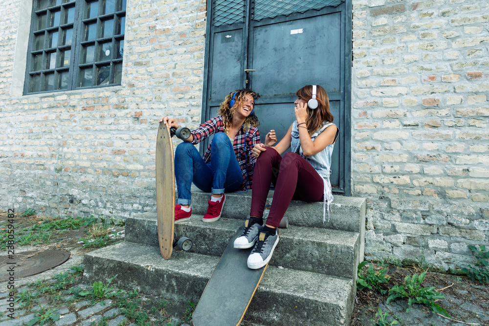 Two girl friends with headphones sitting at the stairs in front of metal door with skateboards and making fun