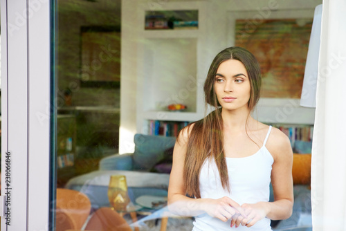 Smiling young woman standing in living room and looking out window