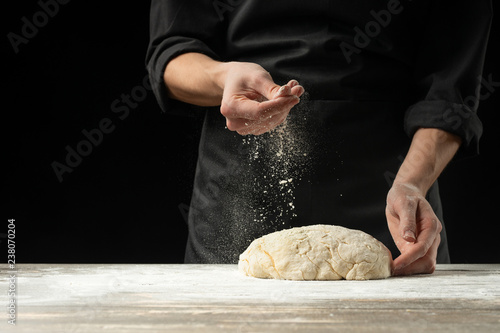 A chef in a black apron on a black background prepares Italian pizza, bread or pasta on a black background. Advertising, design for text, menu, recipe book.