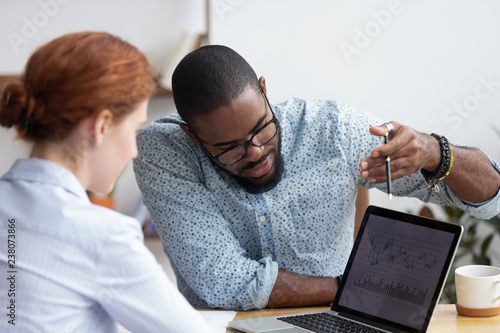 Diverse millennial colleagues working together analyzing diagram looking at computer screen. Black mentor helps female apprentice understand corporate program explaining interface showing on monitor