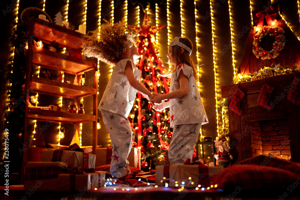 Fototapeta premium Happy little girls wearing Christmas pajamas playing by a fireplace in a cozy dark living room on Christmas eve. Celebrating Xmas at home.