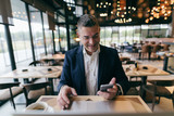 Middle-aged businessman dressed smart casual smiling and using smart phone for call while sitting in cafeteria.