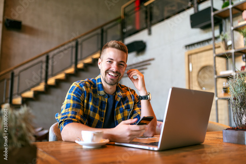 Portrait of young smiling Caucasian freelancer holding smart phone and putting on earphones while sitting in cafe. On the desk laptop and coffee. photo