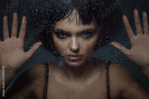Close up portrait of young beautiful woman with provocative make up and stylish bob haircut standing behind the window with rain drops on it and touching the glass with her palms