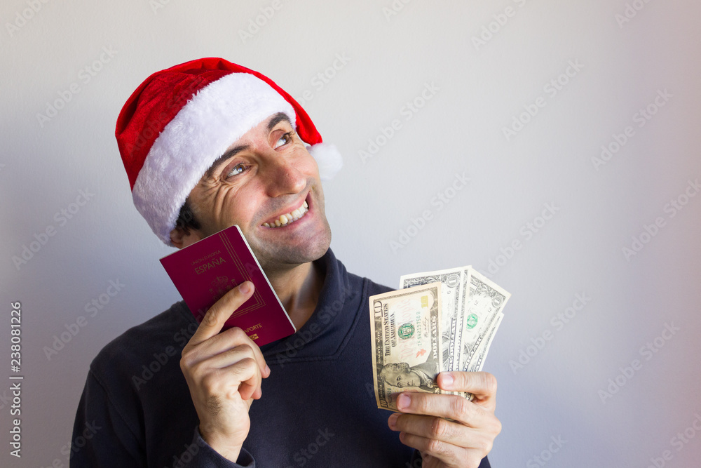 Happy man with Santa hat holding passport and dollar bills over white background. Christmas holidays travel escape plan concept. Language translation: European Union, Spain, passport Happy man with Santa hat holding passport and dollar bills over white background. Christmas holidays travel escape plan concept. Language translation: European Union, Spain, passport